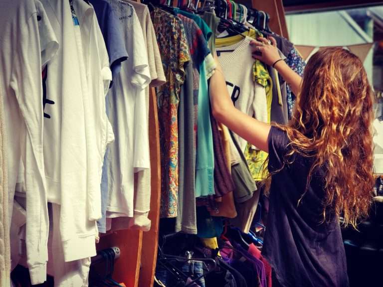 Line of shirts and tops hung on a rack with a long wavy haired person, with their back to the camera, searching through them.